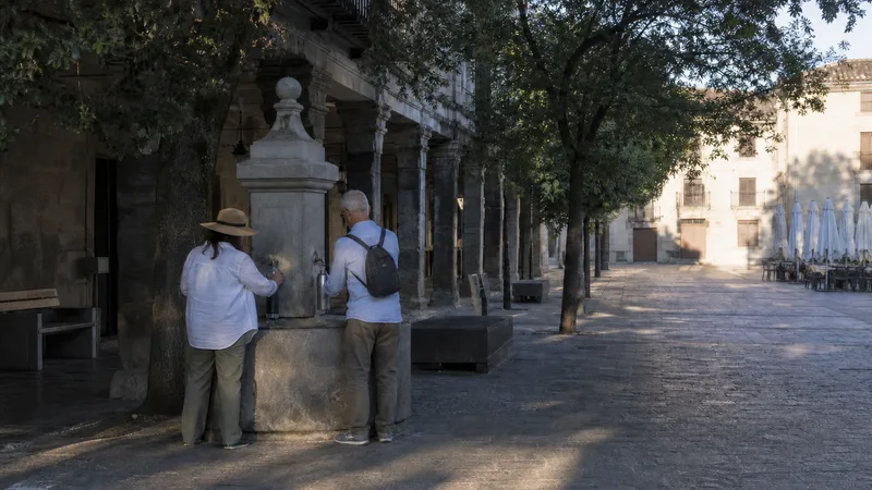 Dos viajeros llenan botellas en una plaza histórica sombreada a primera hora