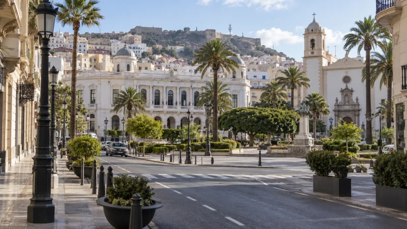 Vista amplia de una plaza urbana de Ceuta, con palmeras, edificios claros y una torre de iglesia.